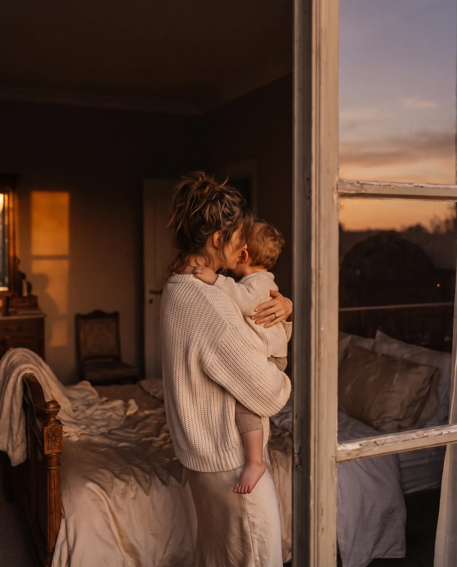 A mother holding her toddler by a window at dusk